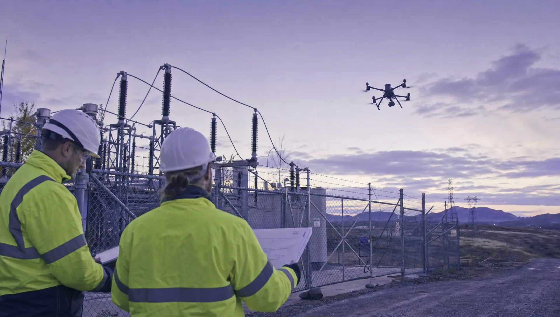 A DJI Matrice 350RTK style drone flies away from a fenced electricity substation while a ground crew studies site plans outside the work zone.