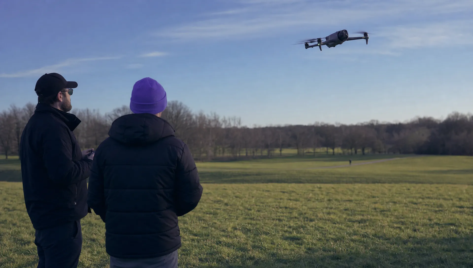 A drone refresher exercise in a large open field, with a DJI Mavic 4 Pro flying away from two stationary crew members above the grass and clear of a distant public footpath.