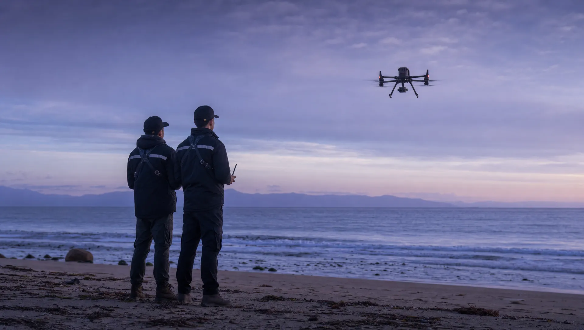 A DJI Matrice 350RTK flies seaward above the shoreline while two crew members stand well back on dry sand.