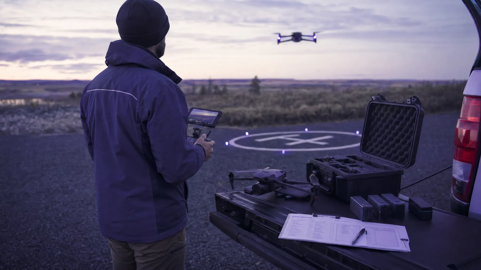 A commercial drone operator conducting a controlled test flight on a marked practice area near an open field edge, with a drone case, spare batteries, controller and printed checklist arranged on a vehicle tailgate beside the launch point.