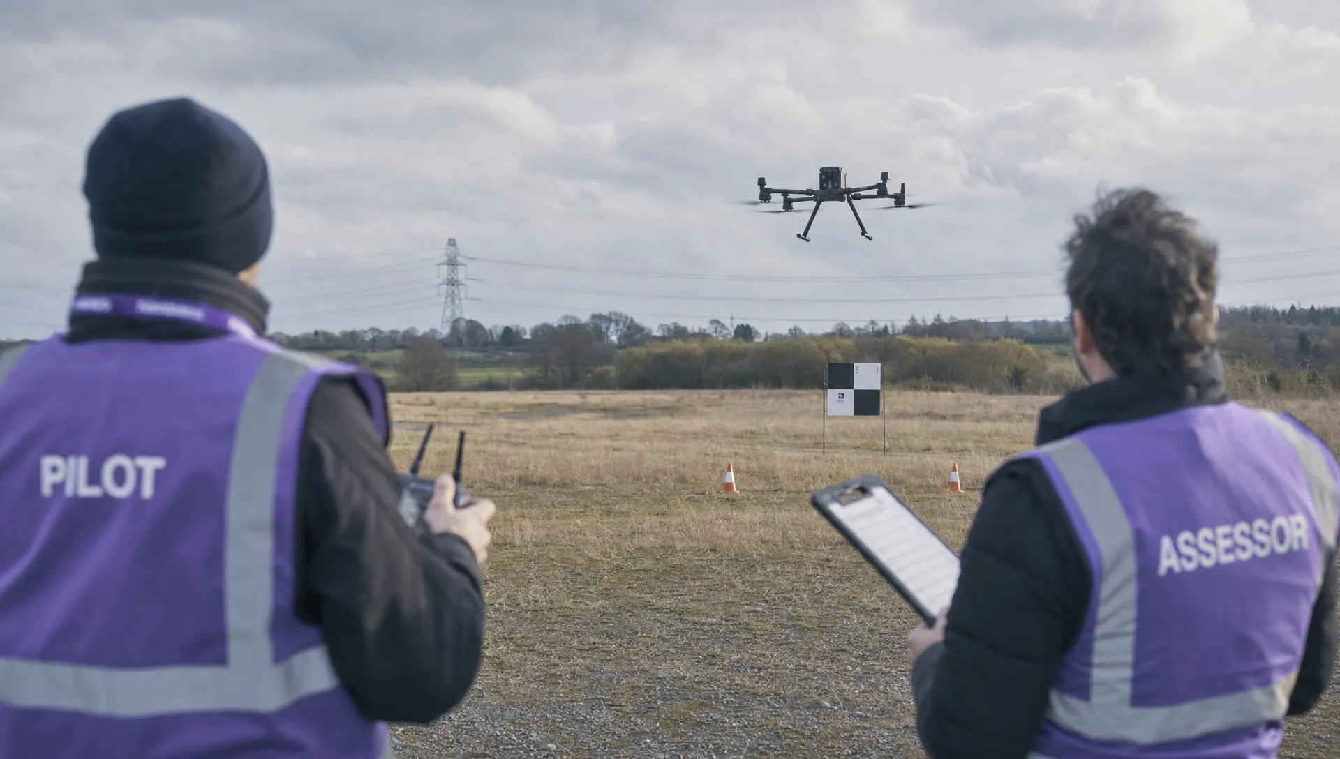 An FAA 107 pilot onboarding assessment in an open training field, with a DJI Matrice 350 RTK flying away from a pilot and assessor toward a mock inspection target.
